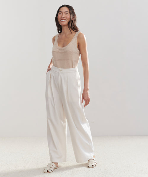 A woman stands smiling, wearing a classic-fit 100% cotton Sweater Tank, high-waisted white pleated trousers, and white sandals against a plain light background.