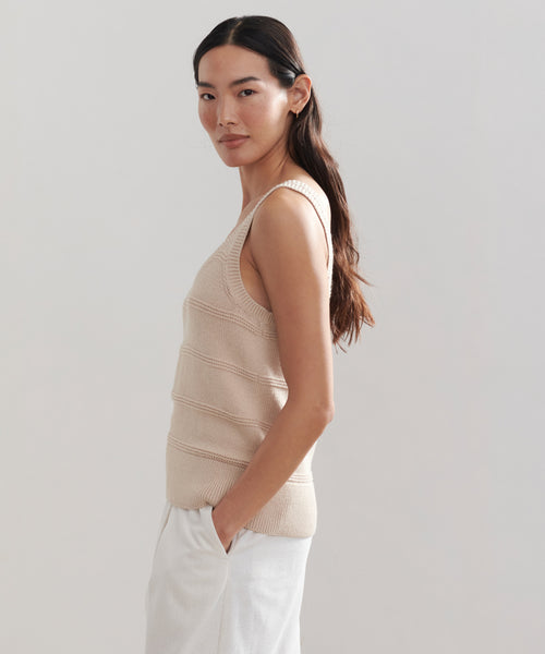 A woman with long dark hair stands in profile against a plain light background, wearing the Sweater Tank and white pants, her hands in her pockets.