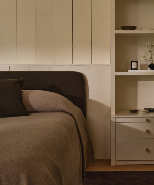 A cozy bedroom corner showcases a Cove Bed with a brown upholstered Libeco linen headboard, matching pillows, and a white shelving unit displaying books, decor, and a flower vase.