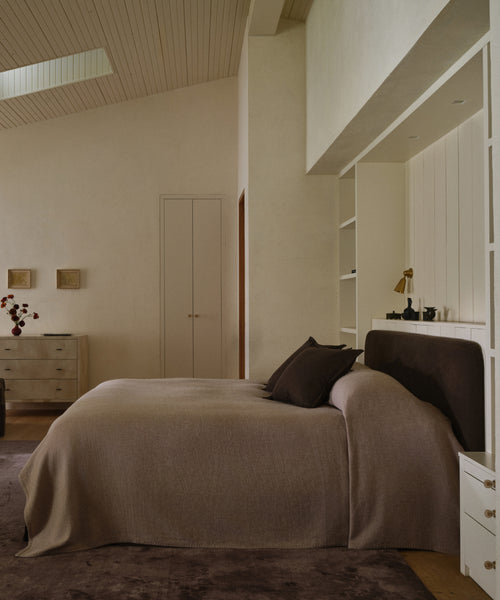 A minimalist bedroom features the Cove Bed with an upholstered headboard, beige bedspread, dark pillows, built-in shelves, a small desk lamp, dresser, soft natural lighting, and simple décor in neutral tones.