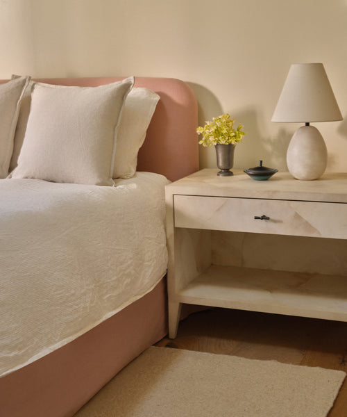 A cozy bedroom with a Cove Bed that has a linen-upholstered headboard, white Belgian Libeco Linen bedding, and a beige nightstand displaying a round white lamp, yellow flowers in a vase, and a dish against cream-colored walls.