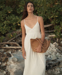 A woman stands outdoors on a rocky beach or shoreline wearing a white sleeveless maxi dress with thin straps and holding a large woven straw tote bag.