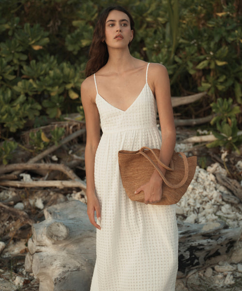 A woman stands outdoors on a rocky beach or shoreline wearing a white sleeveless maxi dress with thin straps and holding a large woven straw tote bag.