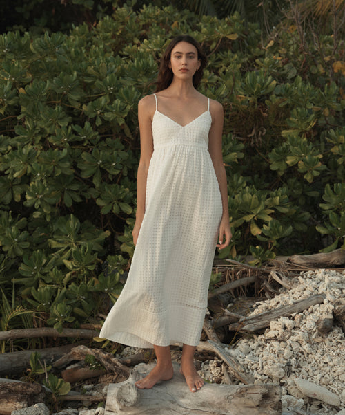 A woman stands barefoot on driftwood in front of dense green foliage, wearing a sleeveless white maxi dress with thin straps and a gathered waist.