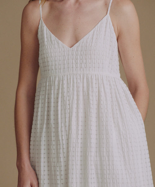 A woman wearing a white sleeveless spaghetti-strap dress with a textured, small-scale geometric pattern, shown against a solid beige background.