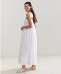 Woman with long brown hair wears the Cove Dress, a sleeveless, relaxed-fit white maxi dress, paired with white sandals. She stands sideways and looks over her shoulder against a plain light background.
