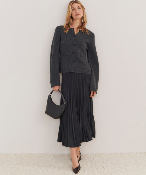 A woman stands against a light wall wearing a dark grey buttoned jacket, black pleated midi skirt, black heels, and carrying the Crescent Shoulder Bag—crafted in Italy from soft leather.