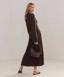 A woman in a brown sweater and pleated skirt stands sideways, holding the Crescent Shoulder Bag—handmade in Italy from soft leather—paired with brown sandals against a plain light background.