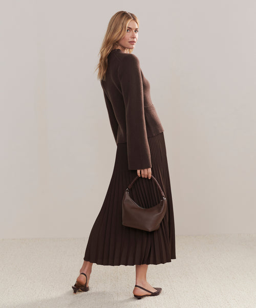 A woman in a brown sweater and pleated skirt stands sideways, holding the Crescent Shoulder Bag—handmade in Italy from soft leather—paired with brown sandals against a plain light background.