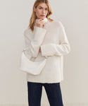 A woman in a cream turtleneck and dark jeans poses with the Crescent Shoulder Bag, crafted in Italy from supple lambskin, stylishly slung over her shoulder against a plain beige backdrop.