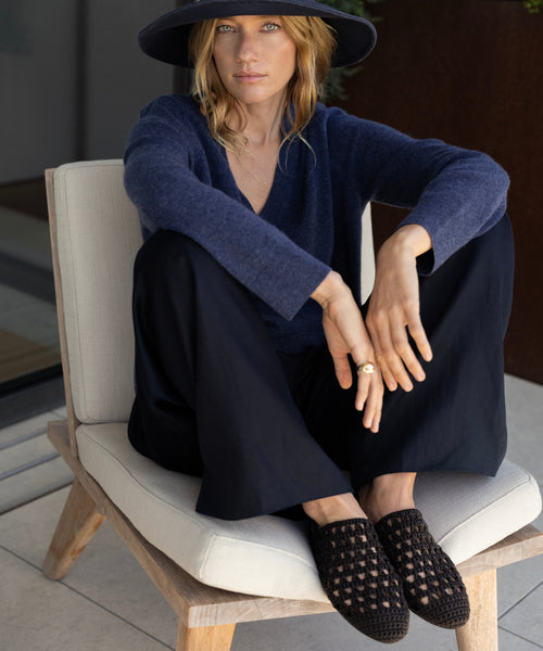 A woman in a wide-brimmed hat, navy sweater, and loose black pants sits cross-legged on a cushioned wooden chair. She gazes calmly at the camera, her feet in Crochet Jules Mules that evoke relaxed Tuscan elegance.