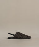 The Crochet Jules Mule, a single black flat-soled open-back shoe, is displayed on a plain beige background, highlighting its hand-crocheted design inspired by the timeless elegance of Tuscany.