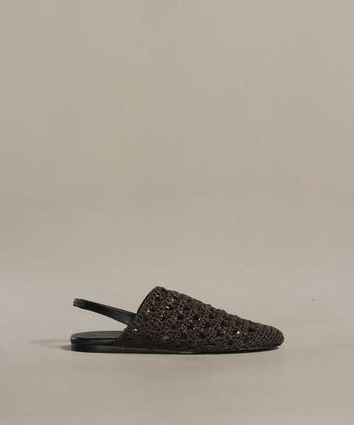The Crochet Jules Mule, a single black flat-soled open-back shoe, is displayed on a plain beige background, highlighting its hand-crocheted design inspired by the timeless elegance of Tuscany.