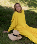A woman in a bright yellow sweater and pleated skirt sits on grass, looking at the camera. Next to her lies a chic Crochet Raffia Sun Hat with a black band.
