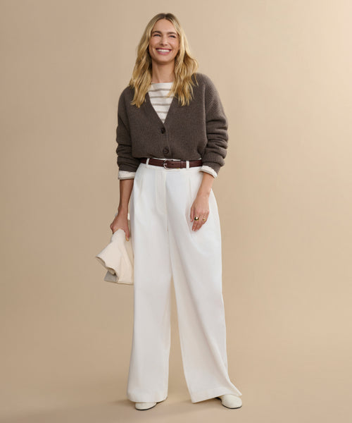 A woman smiles against a beige background, wearing the Cropped Cashmere Cocoon Cardigan over a striped shirt, paired with high-waisted white wide-leg trousers, white shoes, and holding a light-colored clutch.