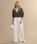 A woman smiles against a beige background, wearing the Cropped Cashmere Cocoon Cardigan over a striped shirt, paired with white wide-leg trousers, white shoes, and holding a light-colored clutch.