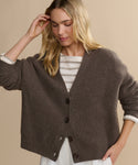 A woman with blonde hair wears the Cropped Cashmere Cocoon Cardigan—an oversized brown button-up—over a white and beige striped top, standing against a beige background, looking down as she touches her hair.
