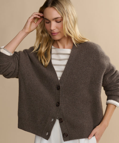 A woman with blonde hair wears the Cropped Cashmere Cocoon Cardigan—an oversized brown button-up—over a white and beige striped top, standing against a beige background, looking down as she touches her hair.