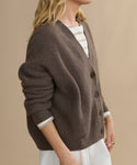 A person with long blonde hair wears the Cropped Cashmere Cocoon Cardigan, featuring large buttons and a ribbed texture, layered over a striped shirt and white pants against a plain beige background.