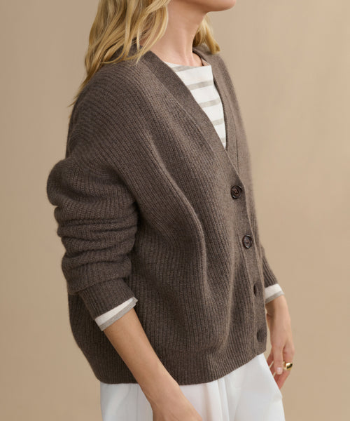 A person with long blonde hair wears the Cropped Cashmere Cocoon Cardigan, featuring large buttons and a ribbed texture, layered over a striped shirt and white pants against a plain beige background.