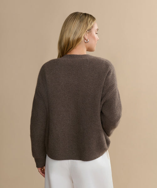 A person with long blonde hair, wearing the Cropped Cashmere Cocoon Cardigan in brown ribbed knit and white pants, stands facing away against a plain beige background.