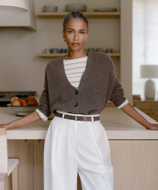 A woman stands in a kitchen wearing the Cropped Cashmere Cocoon Cardigan, effortlessly elevating her everyday look with this versatile piece.
