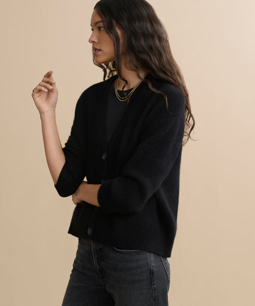 A woman with long wavy hair wears the Cropped Cashmere Cocoon Cardigan over a black top and dark jeans. She stands against a beige background, looking to the side with one arm bent and her hand raised.