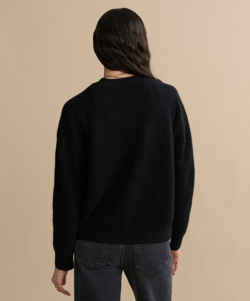 A person with long, wavy dark hair stands with their back to the camera wearing the Cropped Cashmere Cocoon Cardigan in black and dark gray jeans, set against a beige background.