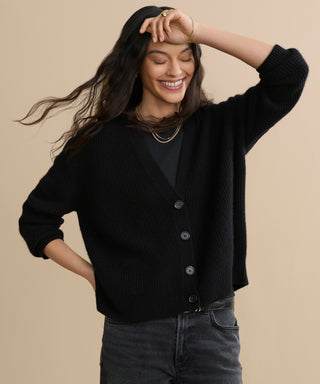 A smiling woman with long dark hair wears the Cropped Cashmere Cocoon Cardigan in black over a matching top and dark jeans, standing against a beige background with one hand on her hip and the other raised above her head.