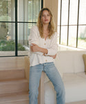 A woman stands indoors by a white sofa, wearing a cream ribbed button-front cardigan and light blue jeans. Large black-framed windows and light wood stairs are visible in the background.