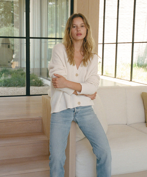A woman stands indoors by a white sofa, wearing a cream ribbed button-front cardigan and light blue jeans. Large black-framed windows and light wood stairs are visible in the background.