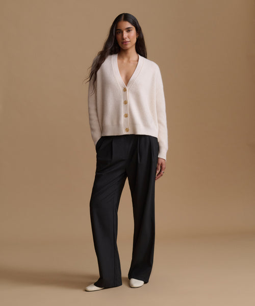 A woman stands against a warm beige studio background wearing a cream cardigan with tan buttons and black wide-leg trousers.