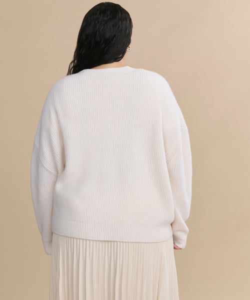 A person with long dark hair is shown from the back, wearing the Cropped Cashmere Cocoon Cardigan and a light pleated skirt against a beige background.