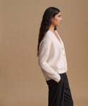 A woman in profile wearing a cream ribbed knit cardigan with buttons and black trousers, standing against a solid beige studio background.