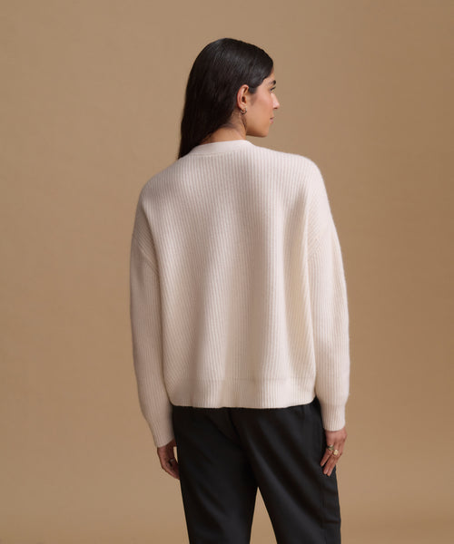 A woman shown from the back wearing a cream ribbed knit sweater and black trousers, standing against a solid tan studio background.