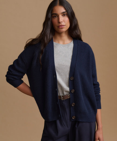 A woman modeling a navy blue ribbed knit cardigan over a light gray crewneck top and navy trousers, set against a solid warm beige studio background.