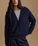 A woman wearing a navy blue ribbed knit cardigan over a light gray top, photographed against a solid warm beige studio background.