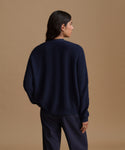 A woman shown from the back wearing a dark navy ribbed knit sweater and matching dark navy trousers, standing against a solid tan studio background.