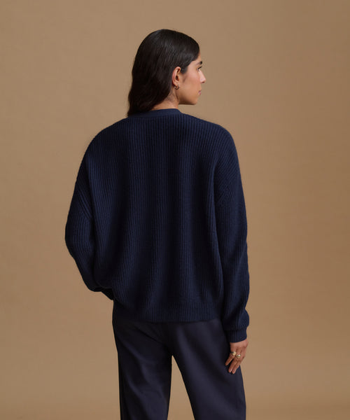 A woman shown from the back wearing a dark navy ribbed knit sweater and matching dark navy trousers, standing against a solid tan studio background.