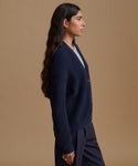 A woman in profile wearing a navy blue ribbed knit cardigan with buttons over a white top and navy trousers, set against a solid beige studio background.