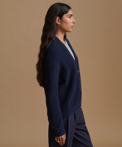 A woman in profile wearing a navy blue ribbed knit cardigan with buttons over a white top and navy trousers, set against a solid beige studio background.