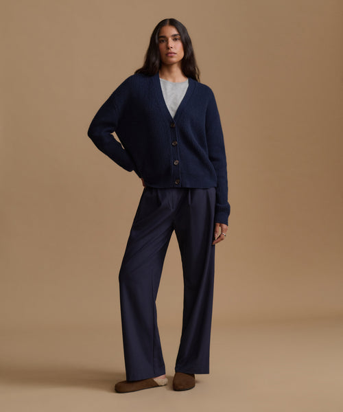 A woman stands against a warm beige studio background wearing a navy blue cardigan and matching wide-leg trousers over a light gray top and brown slip-on shoes.