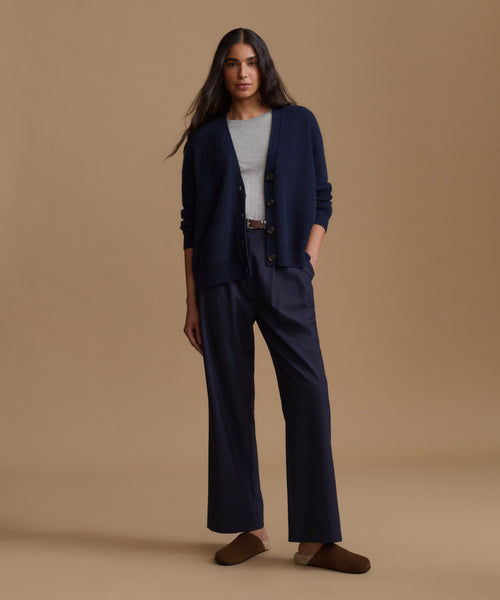 A woman stands against a warm beige studio background wearing a navy blue cardigan and matching wide-leg trousers over a light gray top, with brown slip-on shoes.