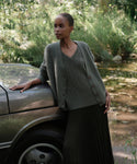 A woman in a Cropped Cashmere Cocoon Cardigan and pleated skirt leans against a car outdoors, surrounded by lush greenery and sunlight.