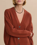 A woman models the Cropped Cashmere Cocoon Cardigan in rust, featuring an oversized ribbed design and large buttons. She pairs it with a black beaded necklace and gold ring, standing against a beige background with her face partially visible.