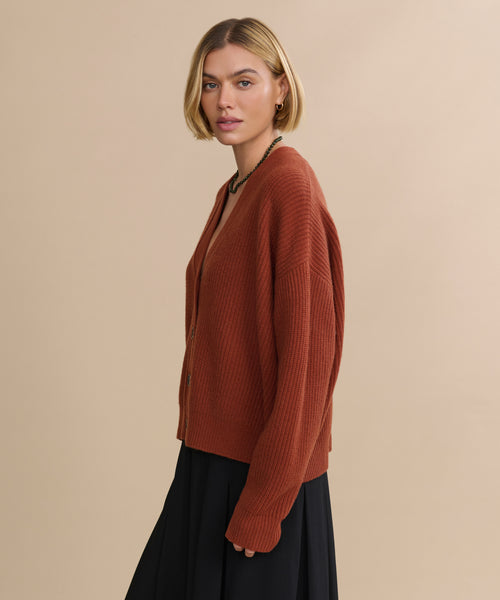 A woman with short blonde hair wears the Cropped Cashmere Cocoon Cardigan in rust and a black skirt, standing against a beige background, facing slightly to the side.