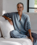 A woman with gray hair tied back, wearing the Cropped Cashmere Cocoon Cardigan and pants, sits cross-legged on a white sofa. Calmly looking at the camera, she rests one arm on the sofa as natural light streams through a window.