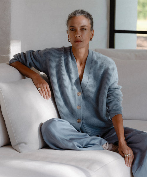A woman with gray hair tied back, wearing the Cropped Cashmere Cocoon Cardigan and pants, sits cross-legged on a white sofa. Calmly looking at the camera, she rests one arm on the sofa as natural light streams through a window.