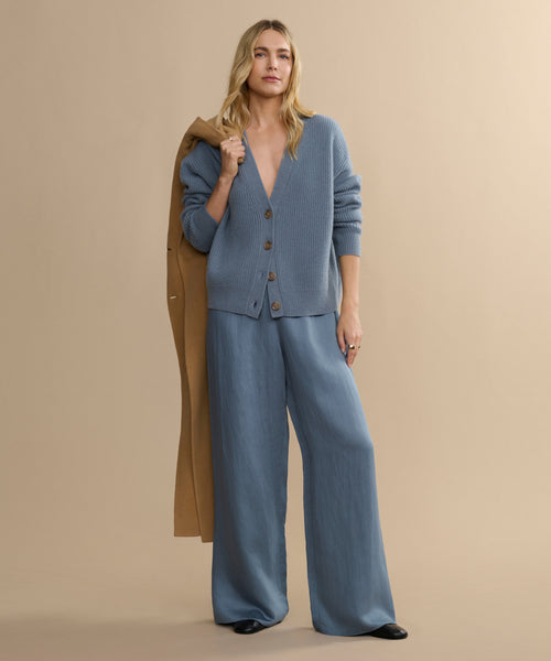 A woman with long blonde hair stands against a beige background wearing the Cropped Cashmere Cocoon Cardigan and wide-leg blue pants, holding a tan coat over her shoulder. She looks relaxed and confident.