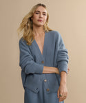 A woman with long blonde hair wears the Cropped Cashmere Cocoon Cardigan and matching pants, standing against a plain beige background and looking confidently at the camera.